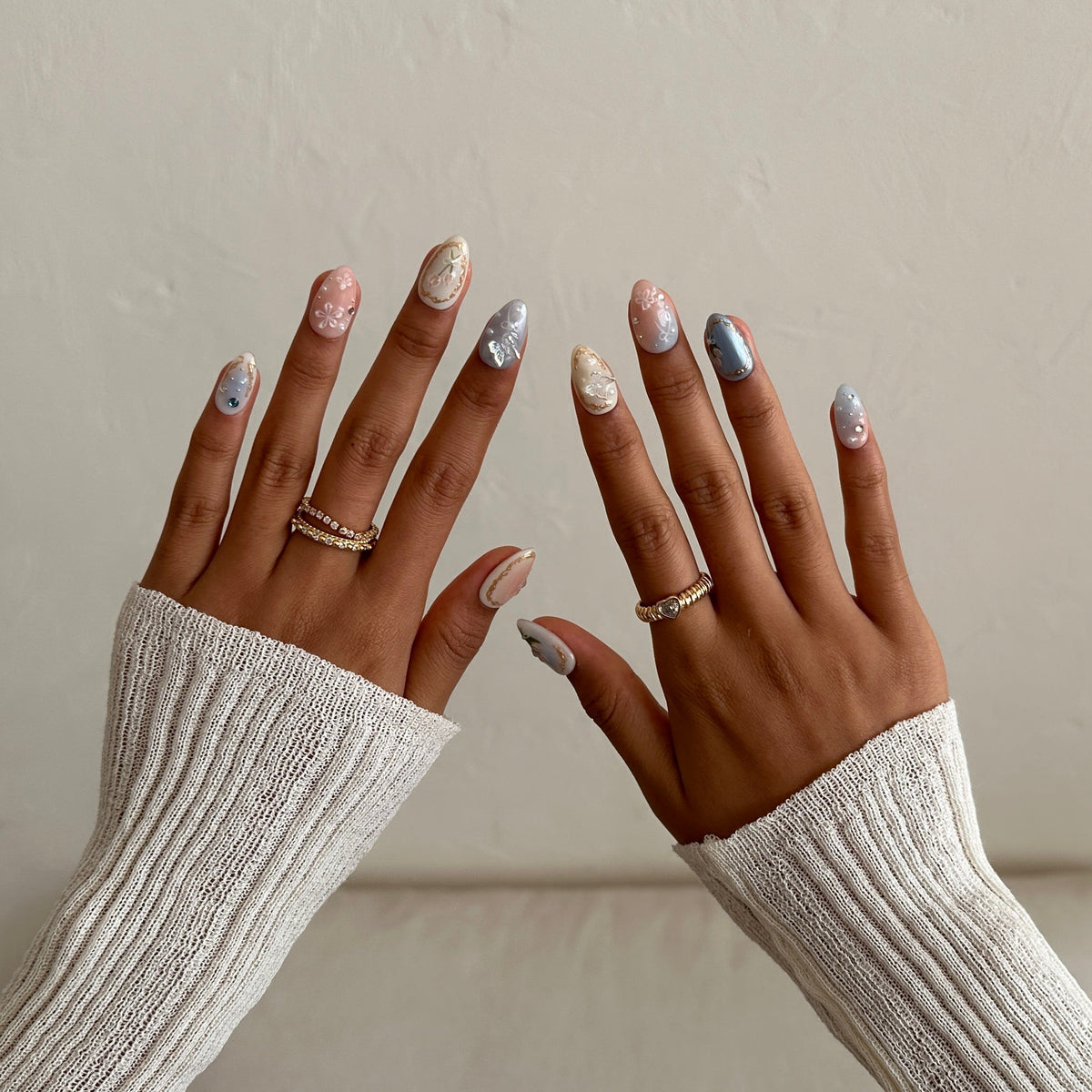 A person's hands with manicured nails painted in pastel shades of pink, blue, and gold, wearing silver rings, shown against a neutral beige wall and sofa background.